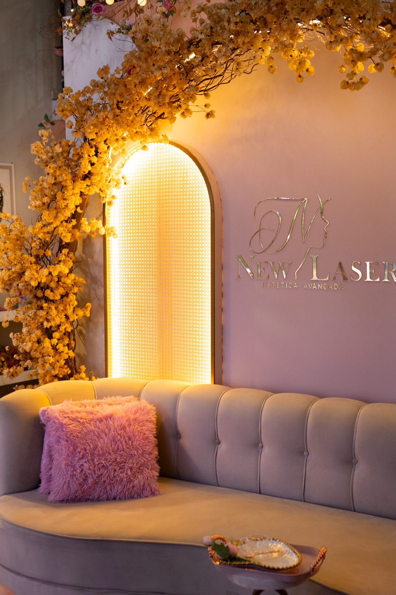 Interior da clínica New Laser Estética Avançada em Toledo/PR, com sofá cinza, almofada rosa, painel luminoso em arco com flores amarelas e logo na parede